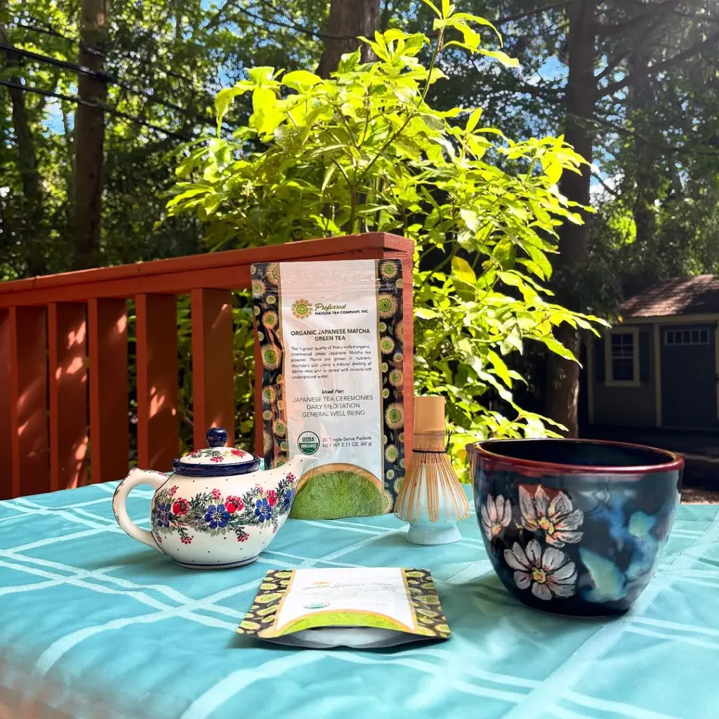 Matcha packages, floral teapot, bamboo whisk, and ceramic cup on an outdoor table