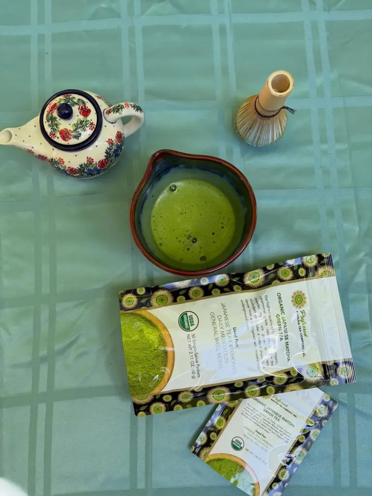 Bowl of green matcha with a floral teapot, bamboo whisk, and Preferred matcha packages