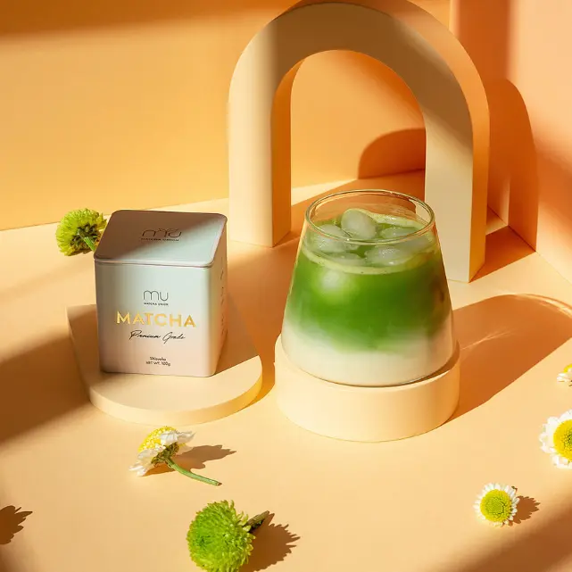 Matcha Union Premium Matcha tin beside a layered iced matcha latte on an orange set with flowers