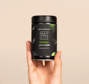 Hand holding a black Matcha & CO Premium Matcha Tea tin against a beige background