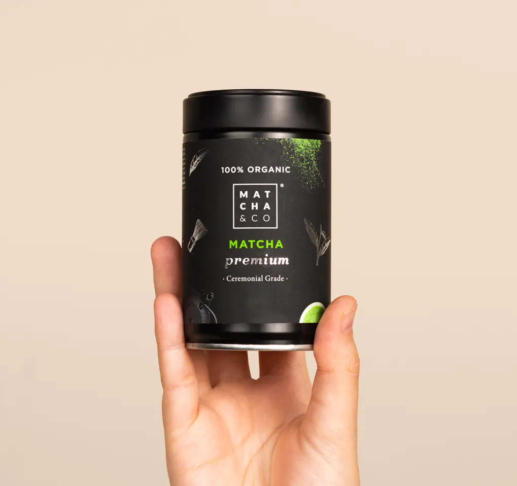 Hand holding a black Matcha & CO Premium Matcha Tea tin against a beige background