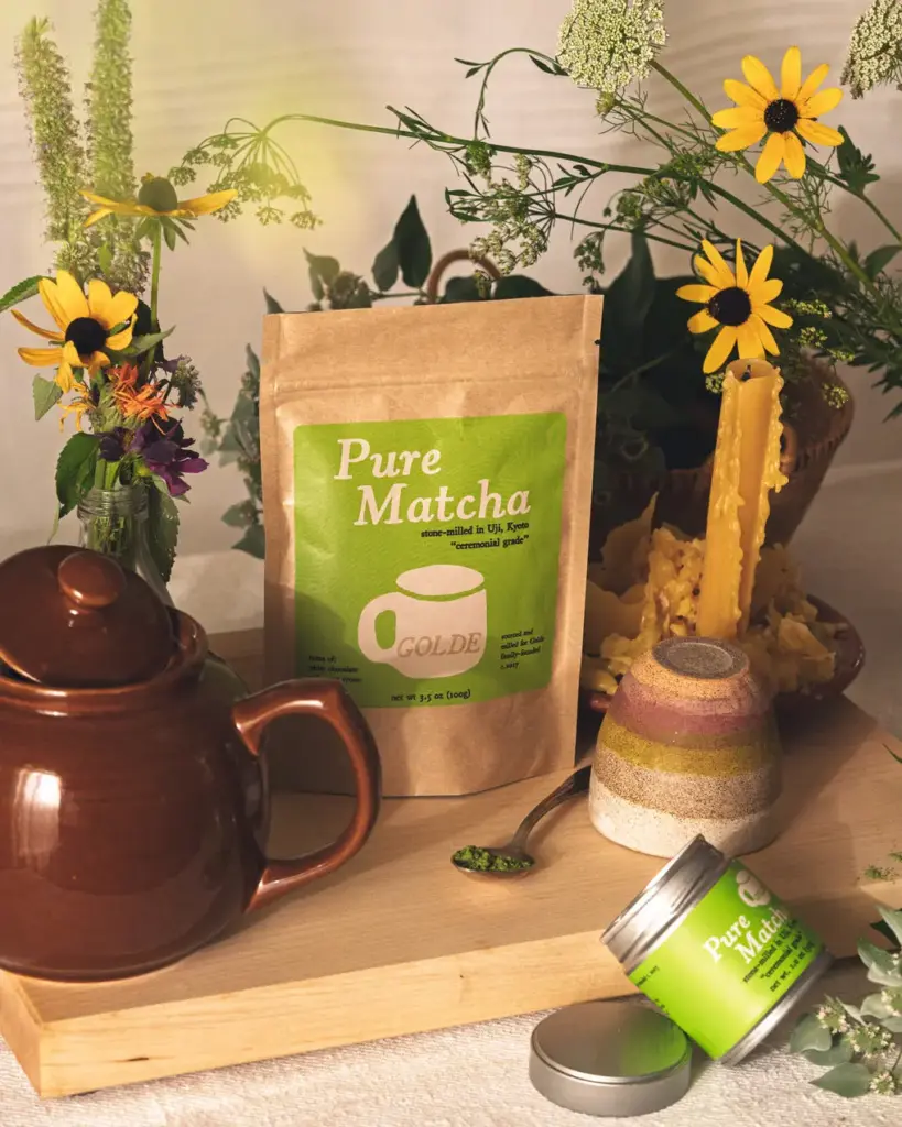 Golde Pure Matcha pouch and tin arranged with a brown teapot, flowers, candle, and spoonful of green powder