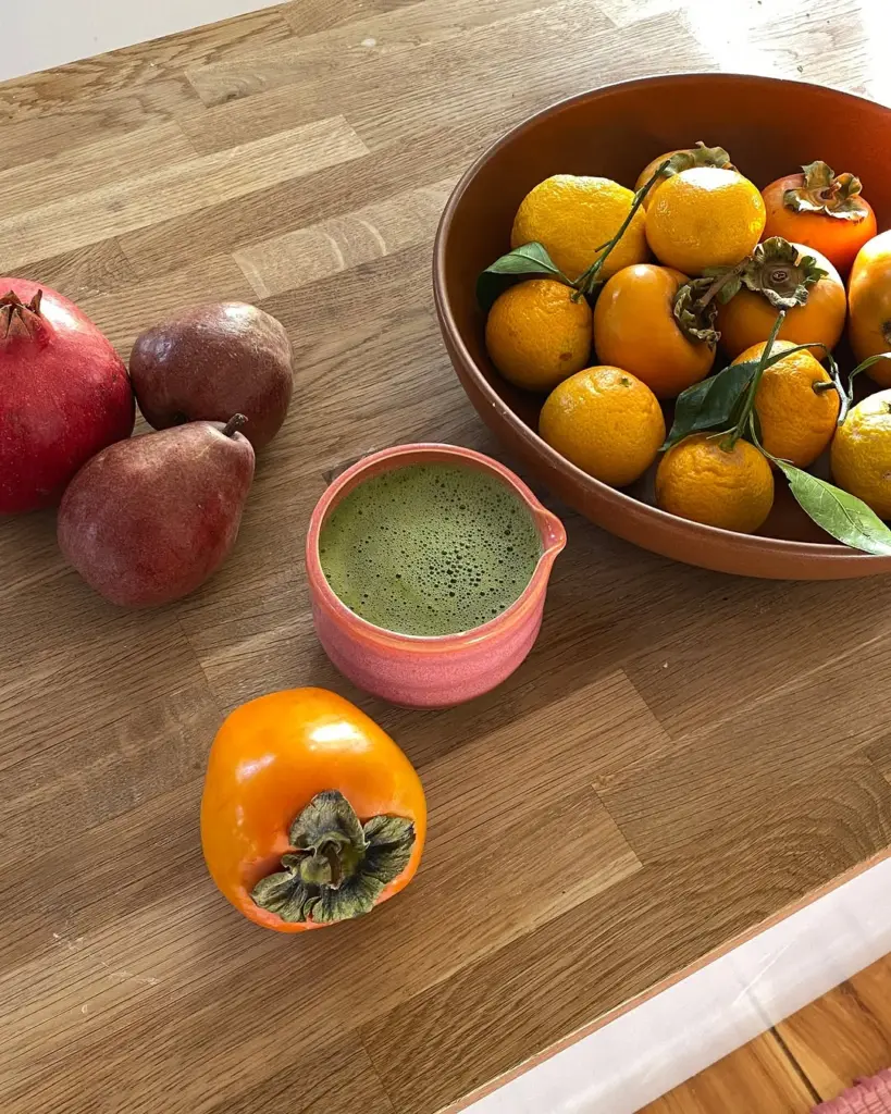 Foamy green matcha in a pink cup beside persimmons, citrus, pears, and a pomegranate