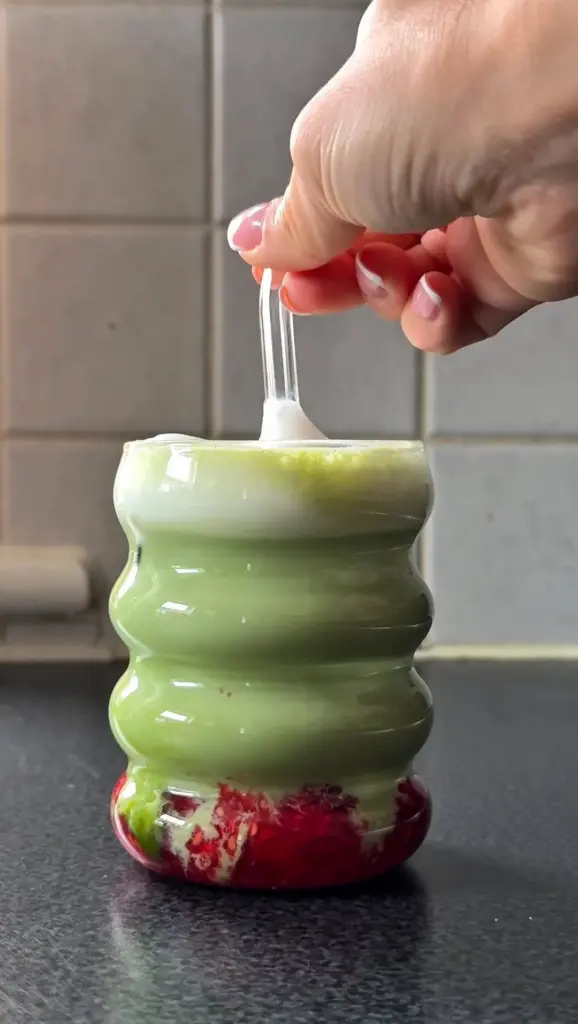 Hand stirring a layered green drink with red berries at the bottom