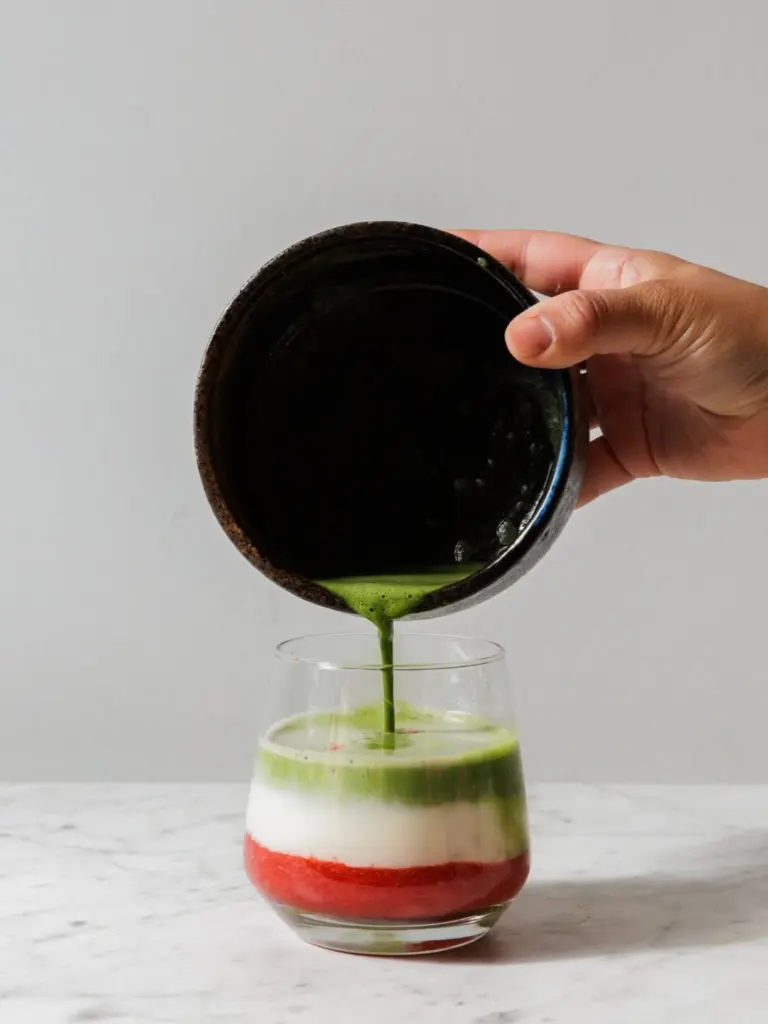 Hand pouring green matcha into a glass with white and red layers