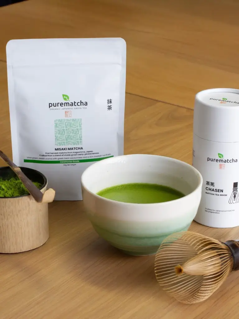 Green matcha in a ceramic bowl beside a bamboo whisk, matcha powder, and PureMatcha packaging