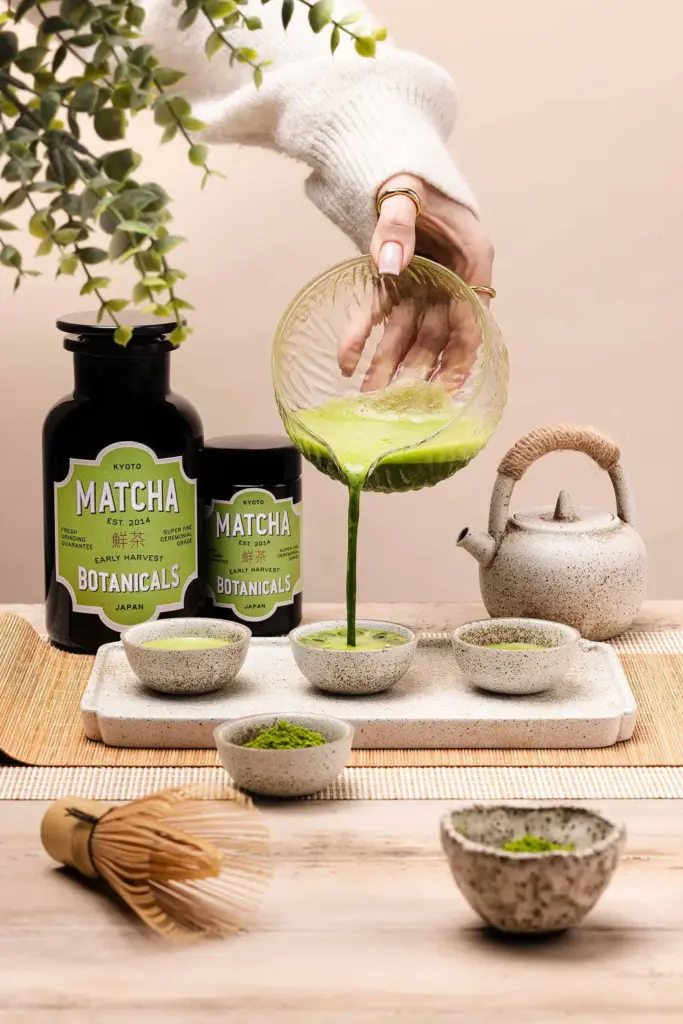 Green matcha poured from a glass bowl into a ceramic cup beside two black Matcha Botanicals containers