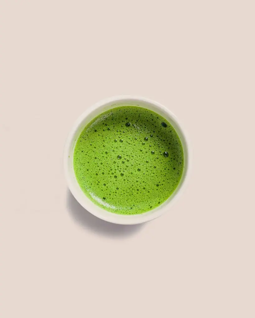 Top view of frothy green matcha in a white cup on a pale beige surface