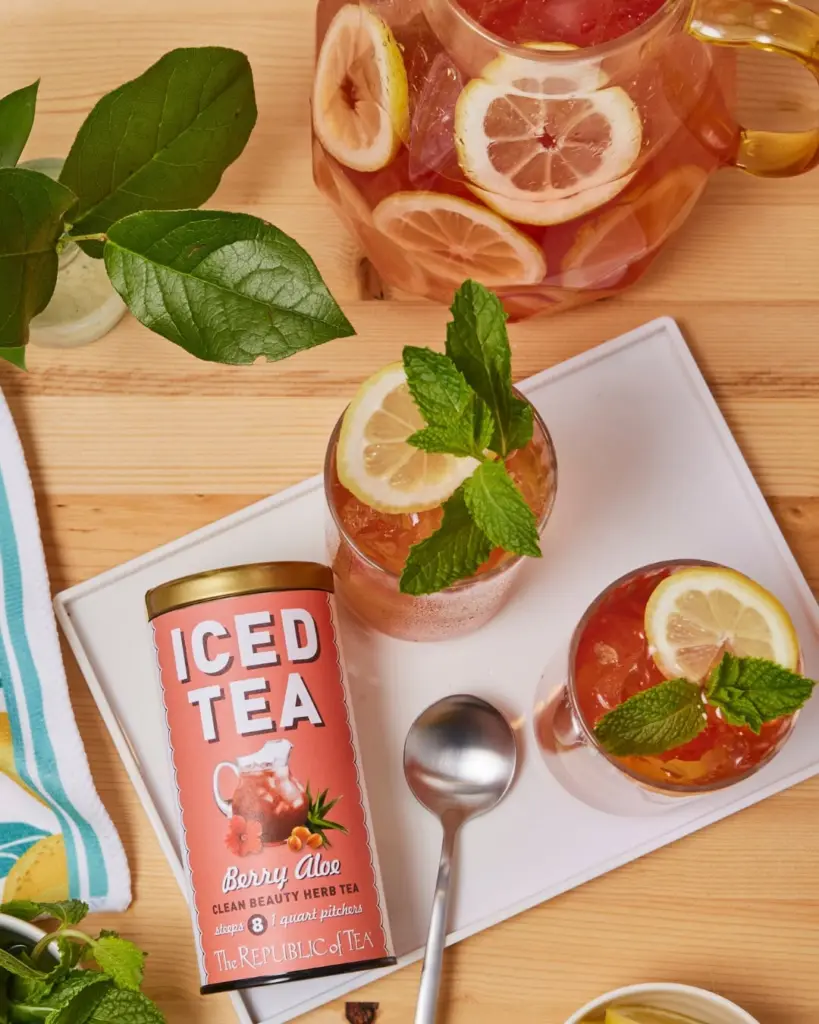 Two glasses and a pitcher of red iced tea garnished with lemon and mint beside a Berry Aloe tin