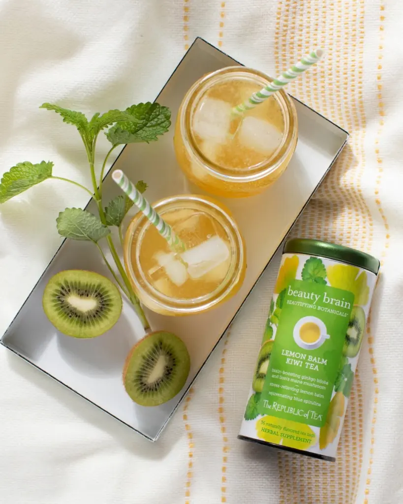 Two glasses of golden iced tea with striped straws, kiwi halves, and a Lemon Balm Kiwi Tea tin
