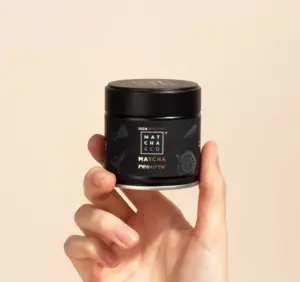 Hand holding a black Matcha & CO Matcha Reserve jar against a pale beige background