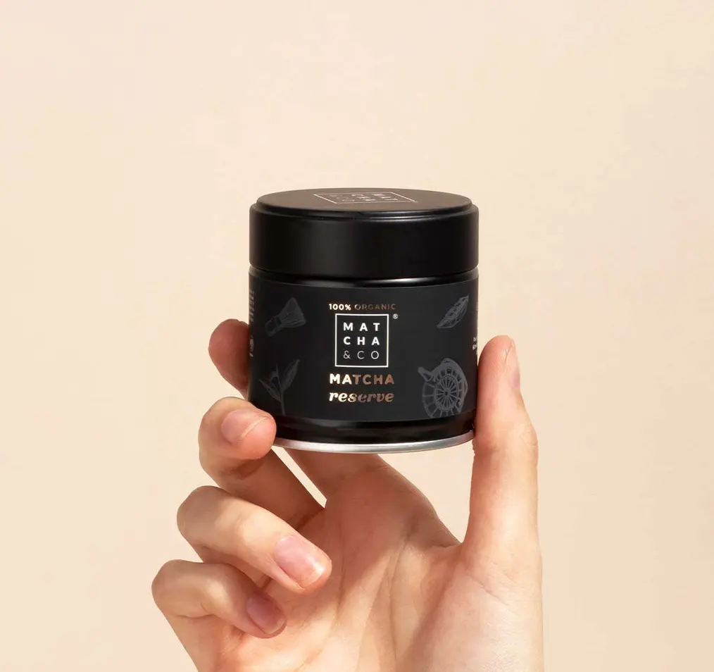 Hand holding a black Matcha & CO Matcha Reserve jar against a pale beige background