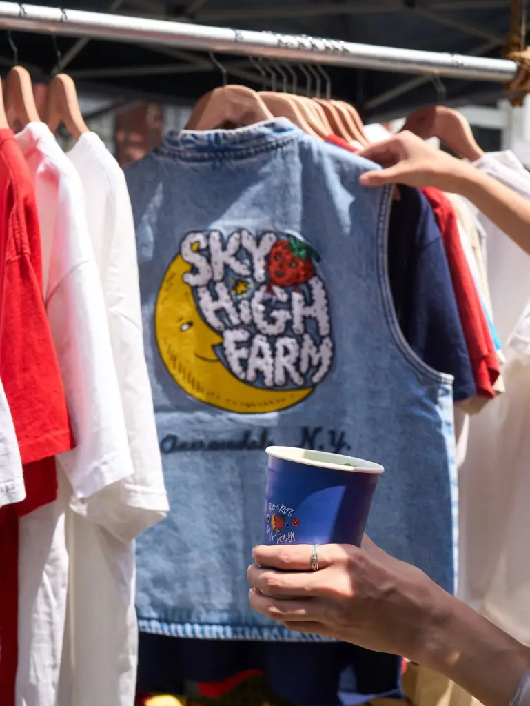 Hands holding a blue cup and a denim vest reading Sky High Farm