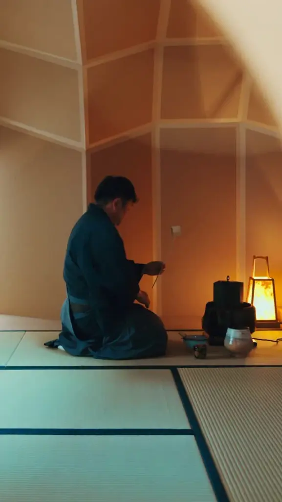Person kneeling on tatami beside tea utensils and a glowing lantern