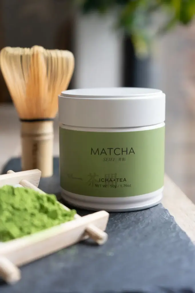 White matcha tin with green label reading Matcha Seiei, ICHA TEA, 50g, beside a bamboo whisk and bright green powder in a wooden scoop on slate