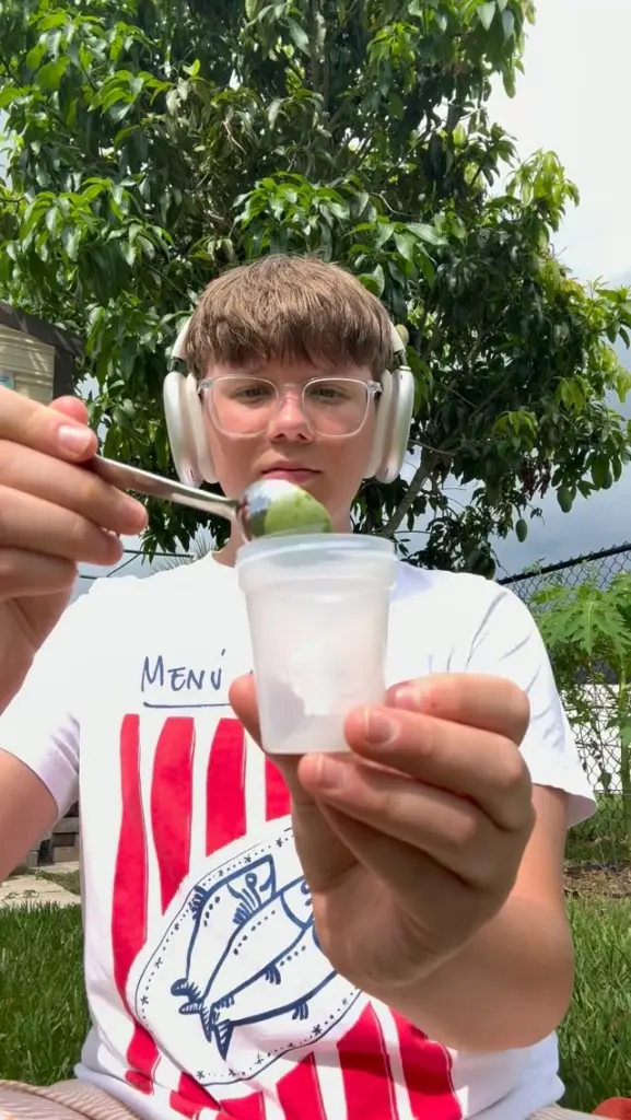 Young person holding a spoonful of green powder over a container outdoors