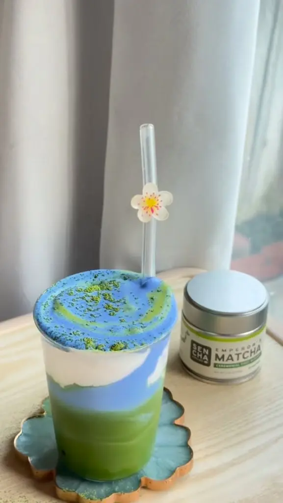 Layered green drink topped with white and blue foam beside a Sencha Naturals matcha tin