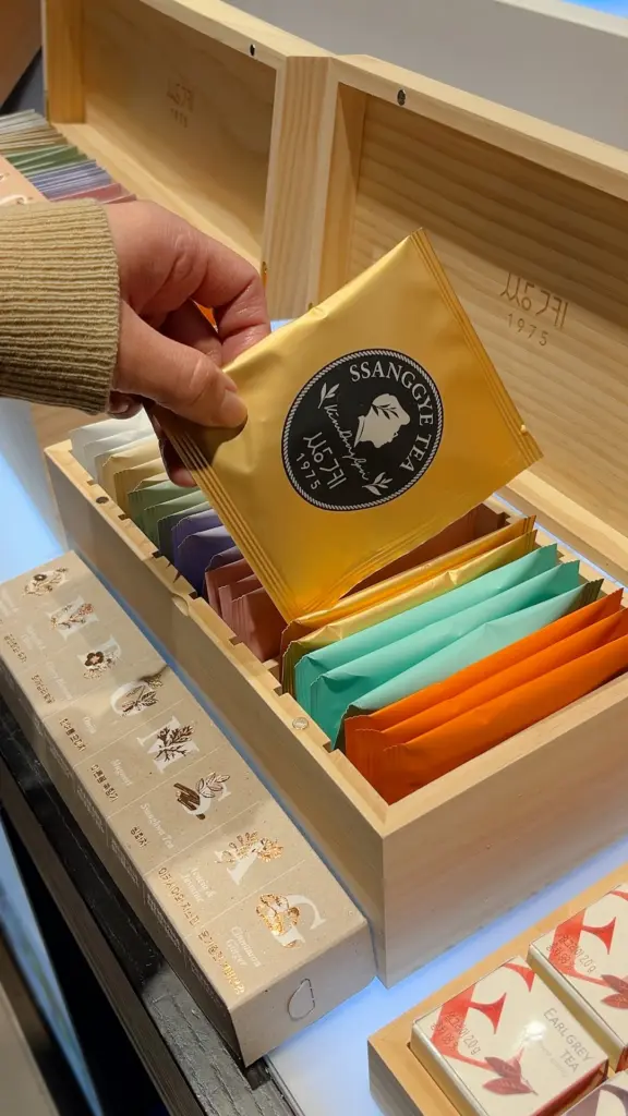 Hand selecting a gold Ssanggye Tea sachet from a wooden box of colorful packets