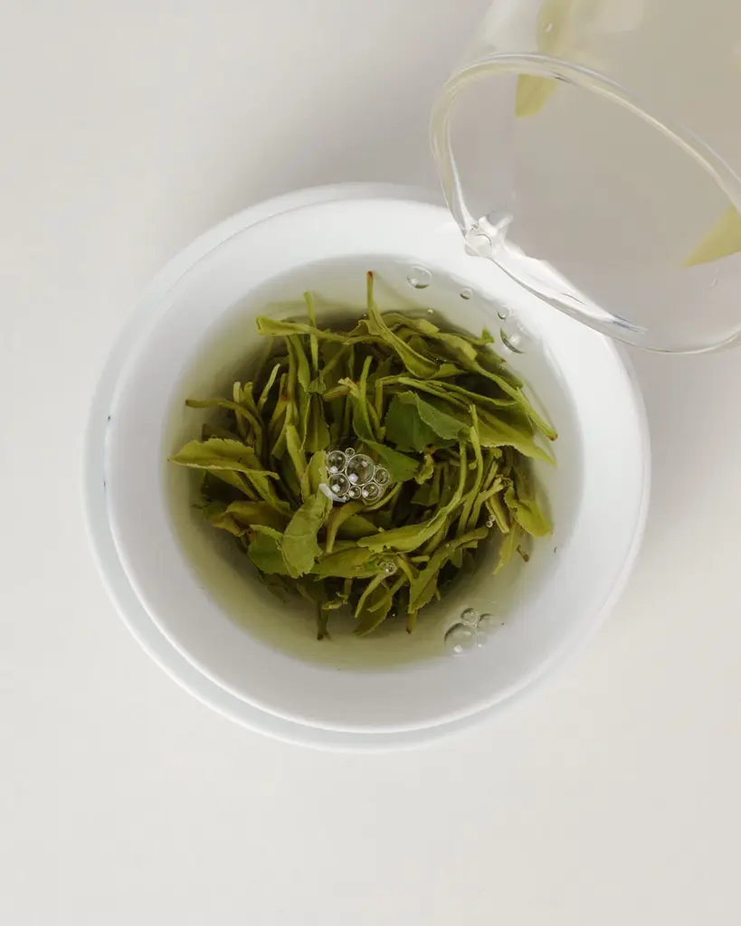 Water pouring over green tea leaves in a white cup
