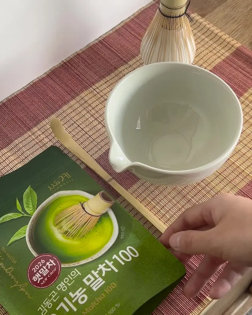 Green tea powder package beside a ceramic bowl, bamboo whisk, scoop, and hand