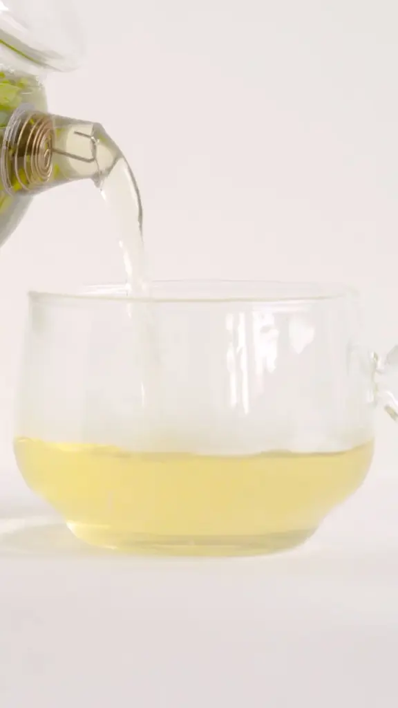 Pale green tea pouring from a glass pitcher into a clear cup