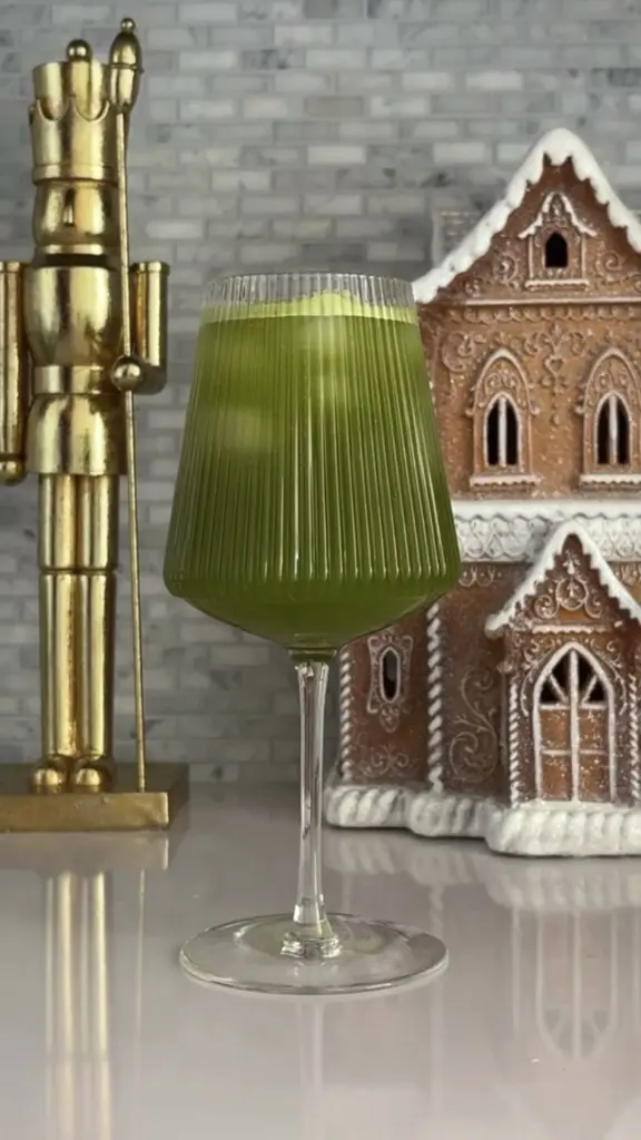 Green frothy drink in a ribbed stemmed glass between a gold nutcracker and gingerbread house