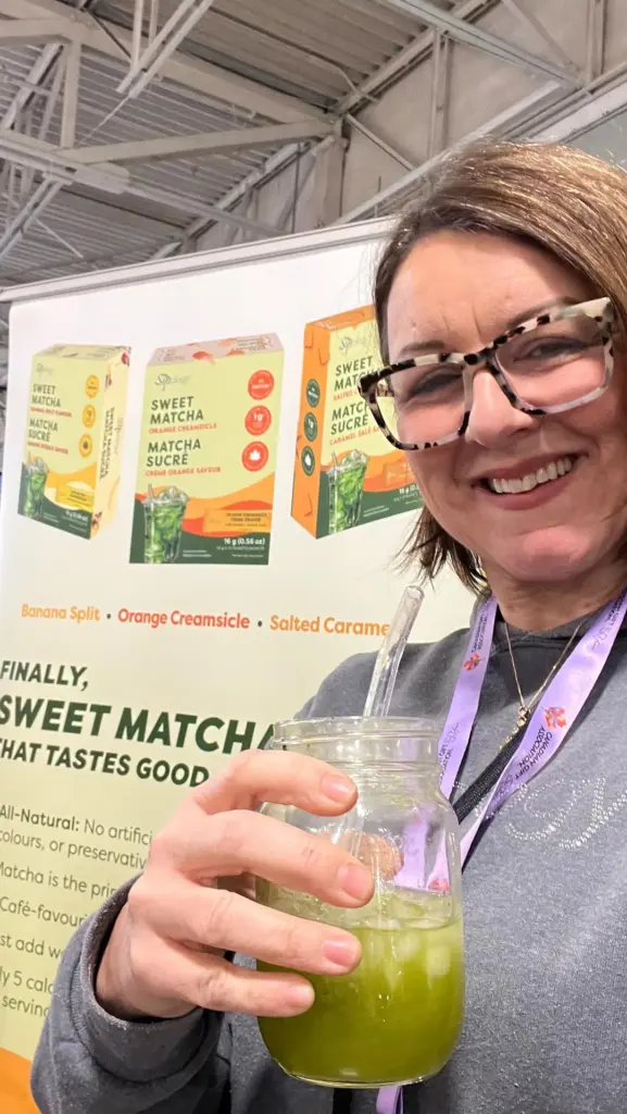 Smiling person holding a mason jar of iced green drink beside a sweet matcha display
