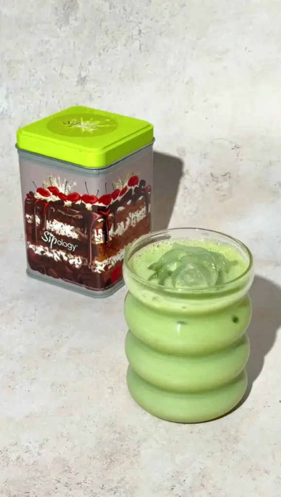 Iced green drink in a ribbed glass beside a colorful Sipology tin