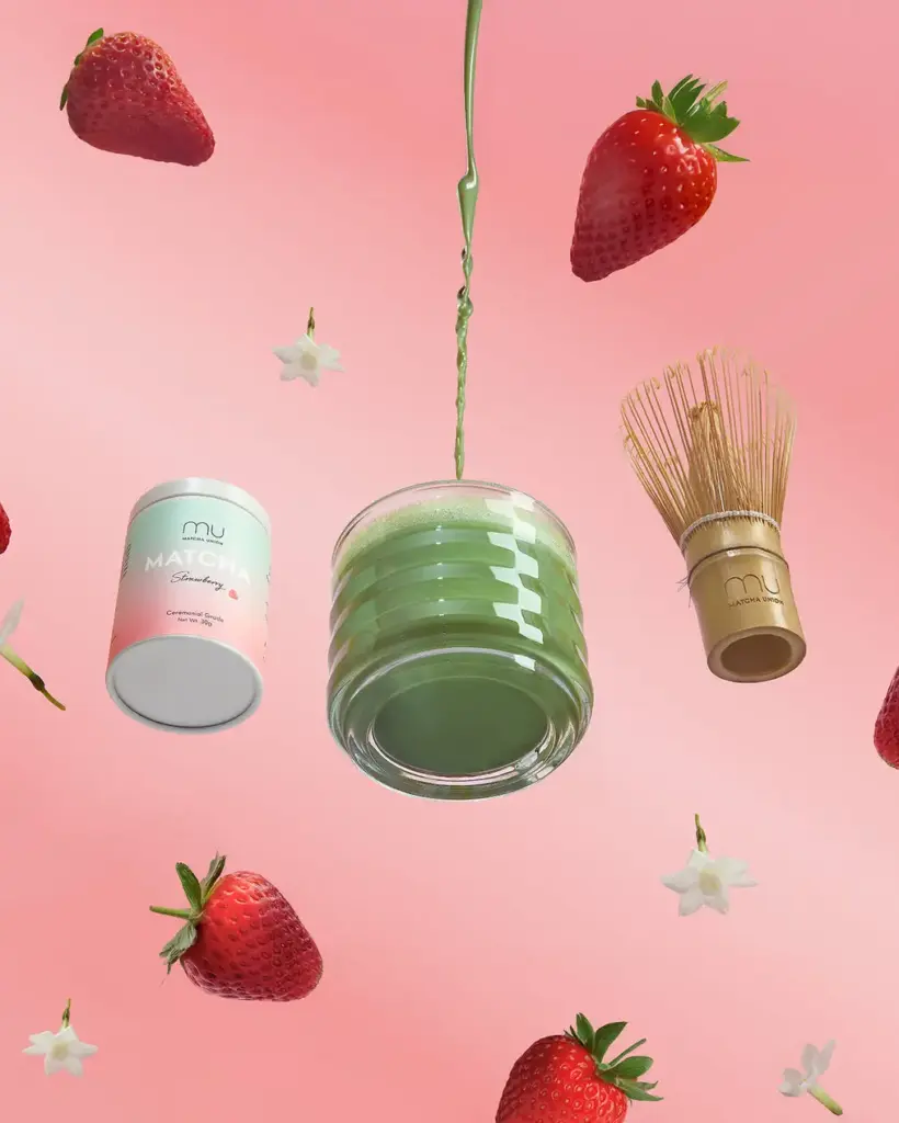 Green matcha drink, Strawberry Matcha tin, bamboo whisk, and strawberries against a pink background