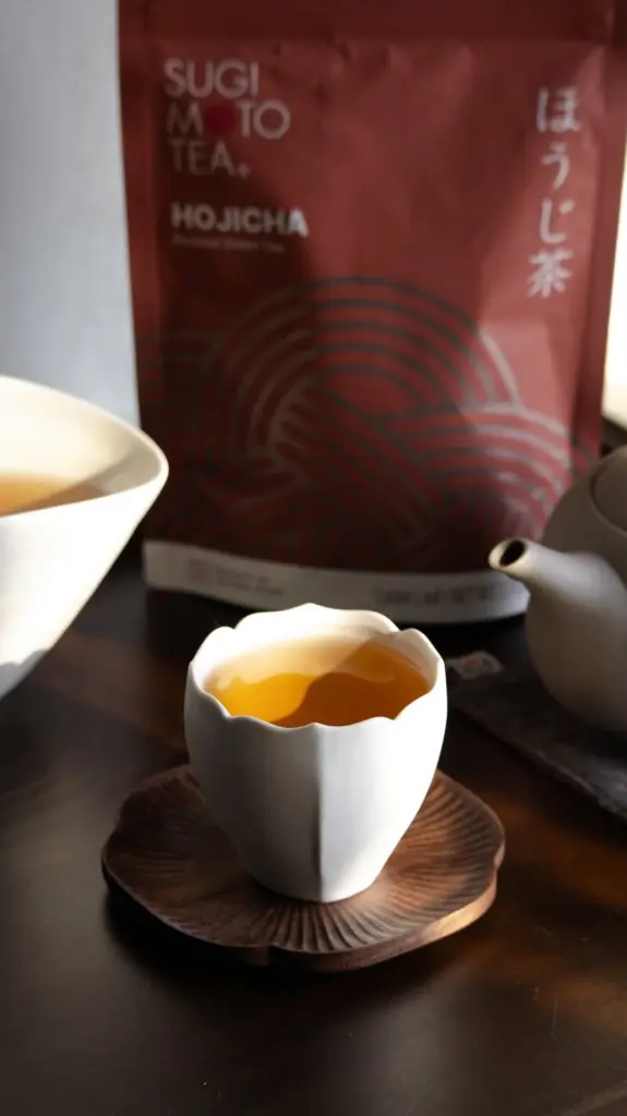 Amber tea in a white ceramic cup beside a teapot and hojicha package