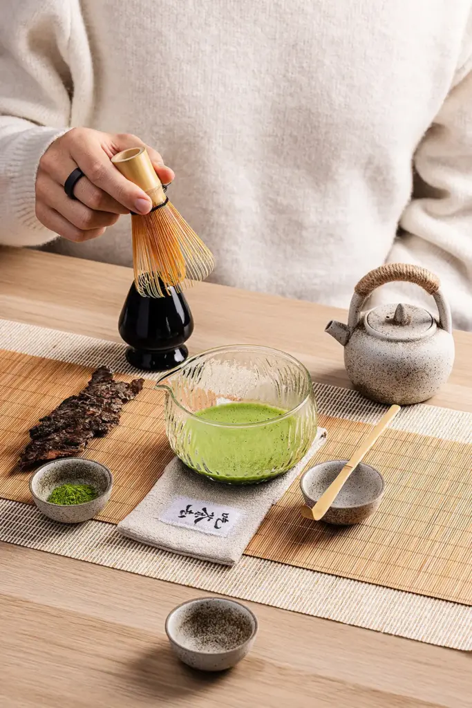 Hand holding a bamboo matcha whisk over a black ceramic whisk stand beside a glass bowl of matcha