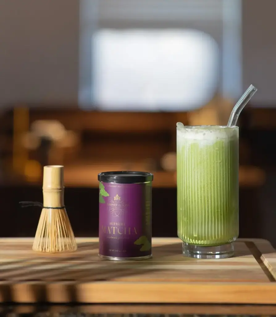 Harney & Sons Supreme Matcha tin beside a bamboo whisk and a tall green drink with foam and a glass straw