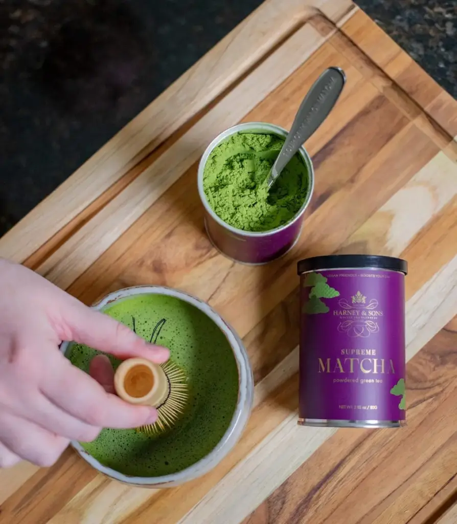 Hand whisking green matcha beside an open powder tin and a purple Harney & Sons Supreme Matcha container