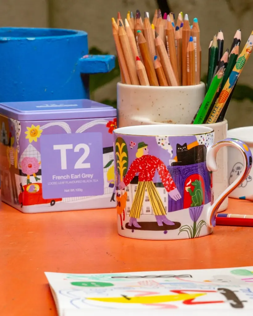 Illustrated mug and French Earl Grey tin among cups of colored pencils