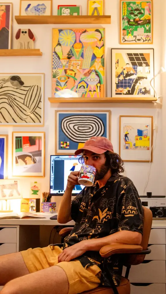 Seated man drinking from an illustrated mug in an art-filled studio