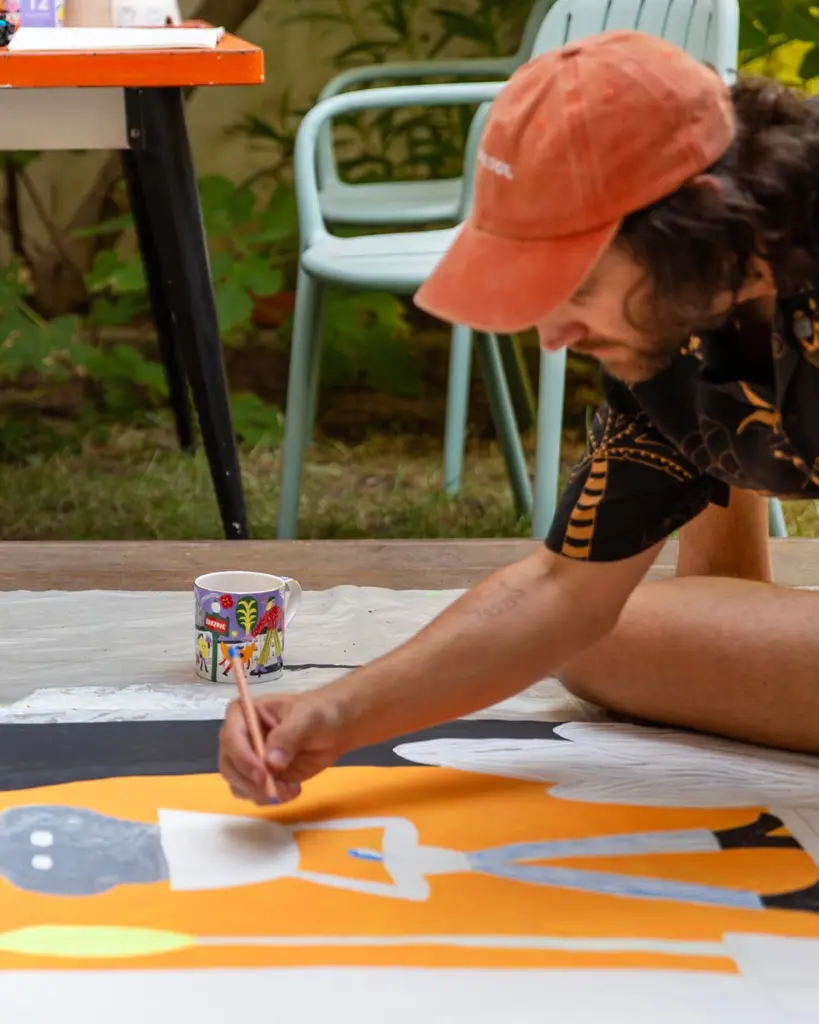 Man painting a large orange artwork beside a colorful mug outdoors