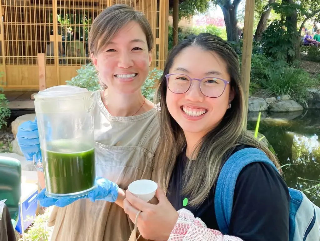 Two smiling women holding a pitcher of green tea and a small white cup
