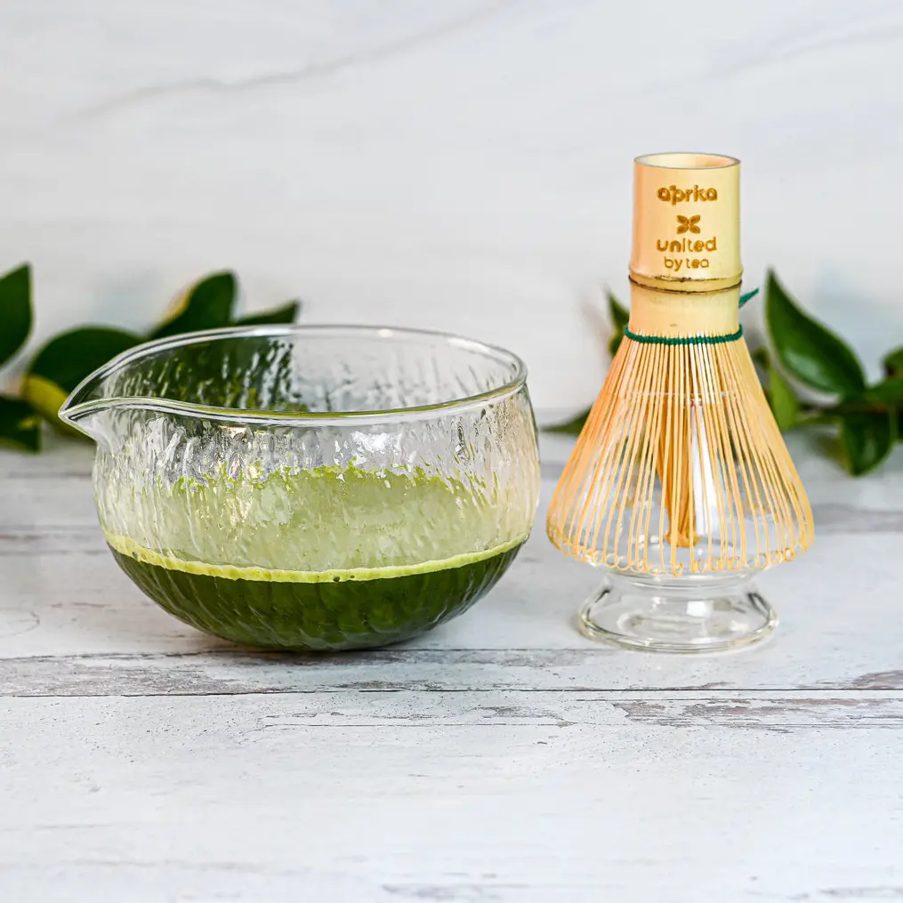 Textured glass bowl with green matcha beside a bamboo whisk on a clear holder