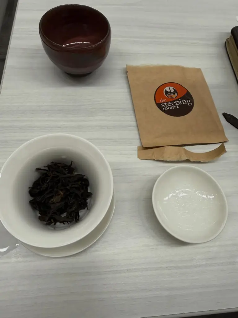 Loose dark tea leaves in a white bowl beside a brown The Steeping Room packet and ceramic bowls