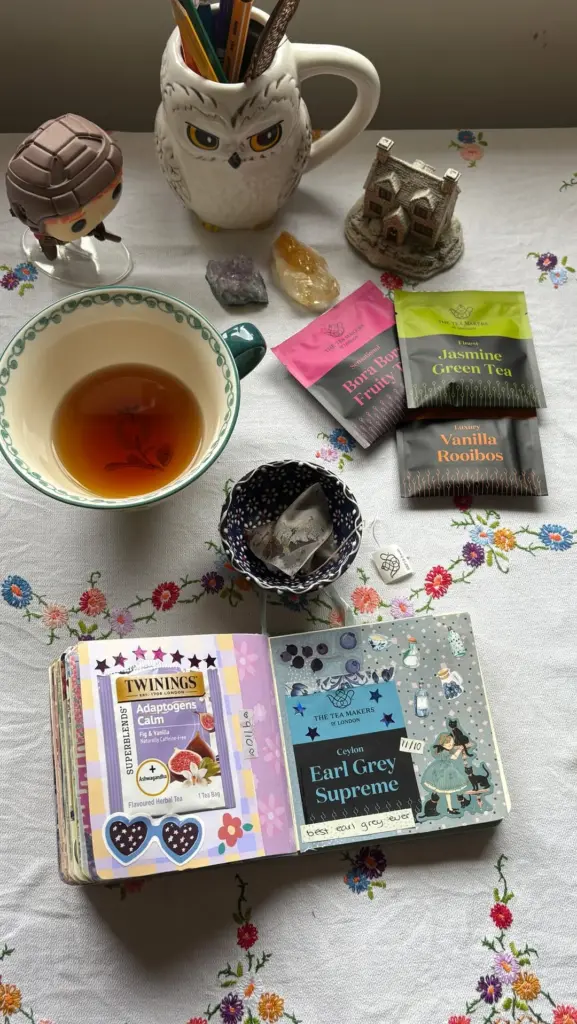 Cup of amber tea beside The Tea Makers packets and an open decorated tea journal