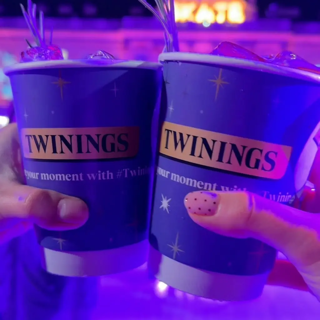 Two hands holding iced drinks in Twinings cups under purple lighting