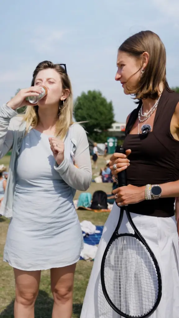 Woman drinking from a Twinings can beside another woman holding a tennis racket and microphone