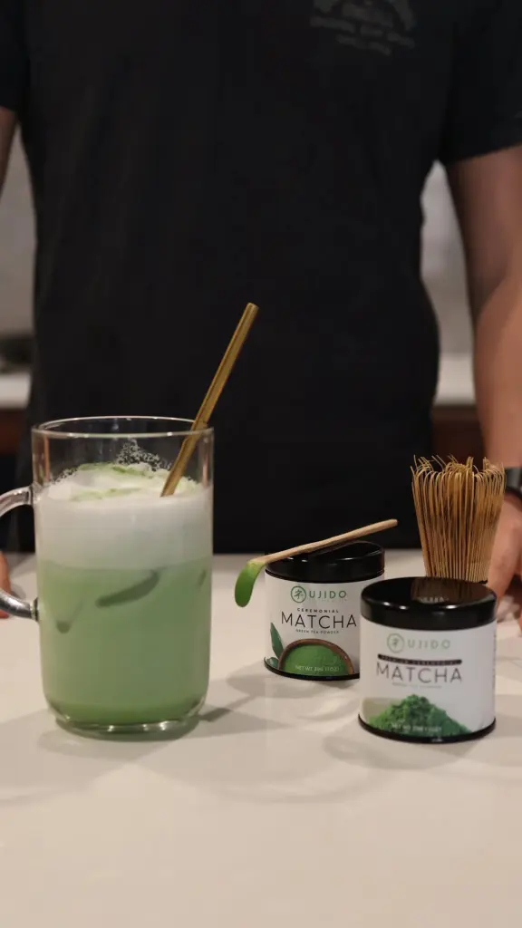 Iced green matcha drink beside two Ujido tea tins, a bamboo whisk, and a powder-filled scoop
