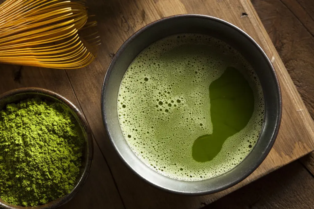 Bowl of frothy green matcha beside matcha powder and a bamboo whisk