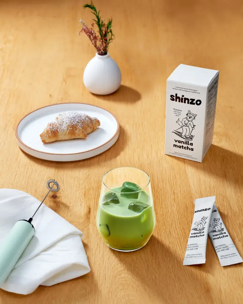 Iced green matcha drink beside Shinzo Vanilla Matcha box and pouches on a wooden table