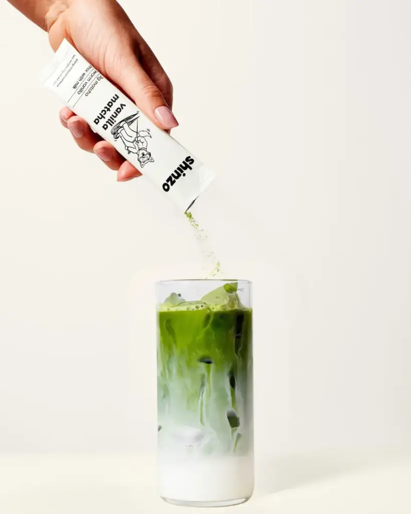 Hand pouring Shinzo Vanilla Matcha powder from a single-serve pouch into an iced milk drink