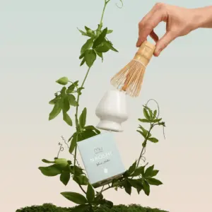 Hand holding a bamboo matcha whisk above a white whisk holder and pale blue Naoshi box