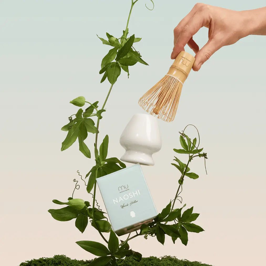 Hand holding a bamboo matcha whisk above a white whisk holder and pale blue Naoshi box