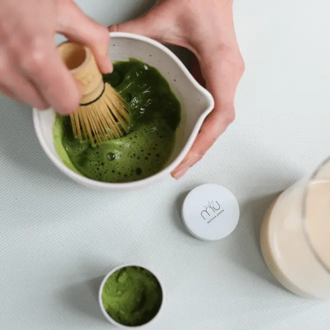 Hands whisking green matcha in a white spouted bowl beside an open tin of powder