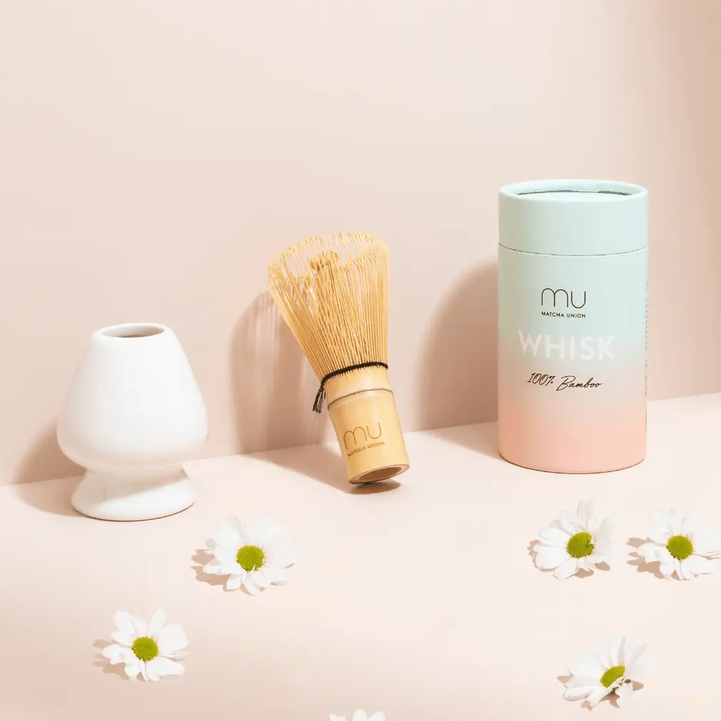 Bamboo Matcha Union whisk beside a white holder and cylindrical package on a pale pink surface